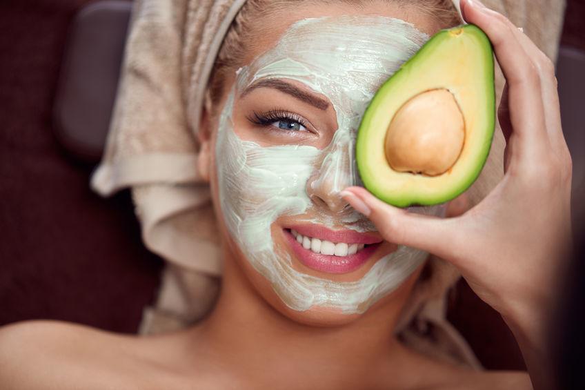 A smiling lady with a cosmetic face mask and holding an avocado over her eye